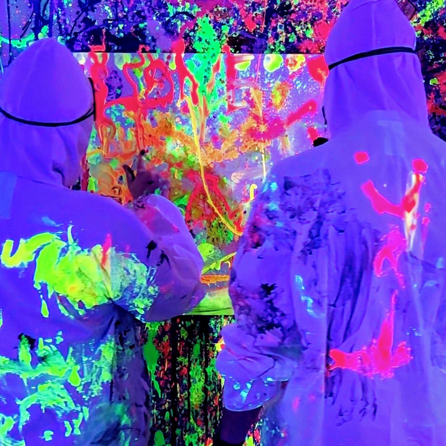 Two people in white suits painting a neon-splattered canvas under UV light.
