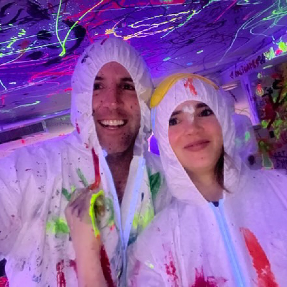 Two people in paint-splattered suits and hoods under blacklight with neon colors.