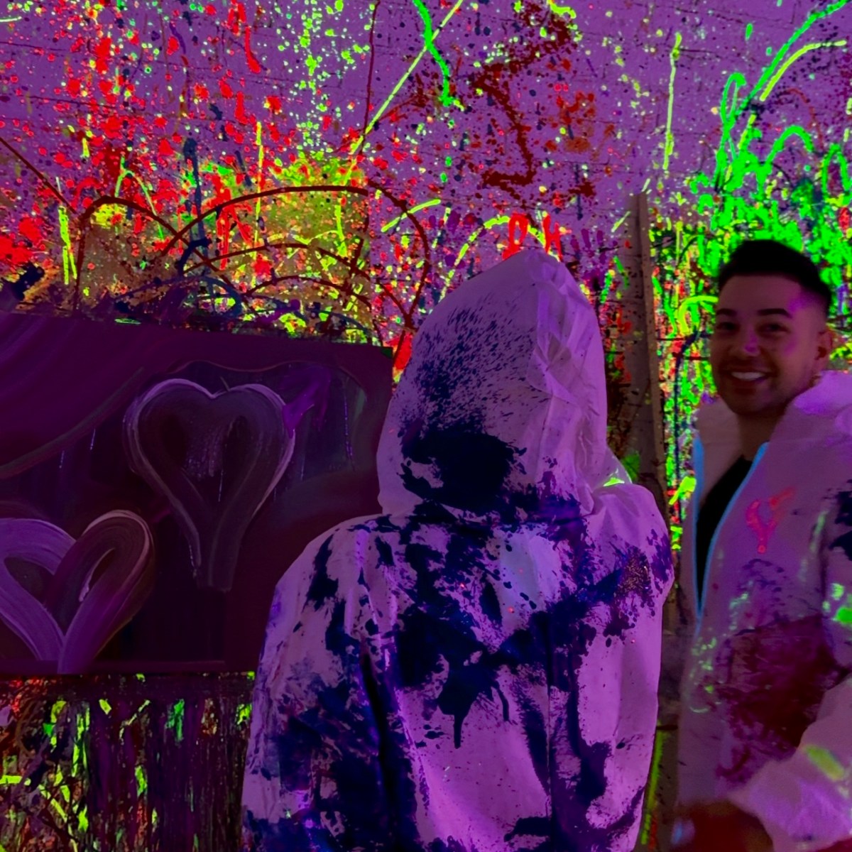 Two people in paint-splattered suits in front of a neon splatter-painted wall.