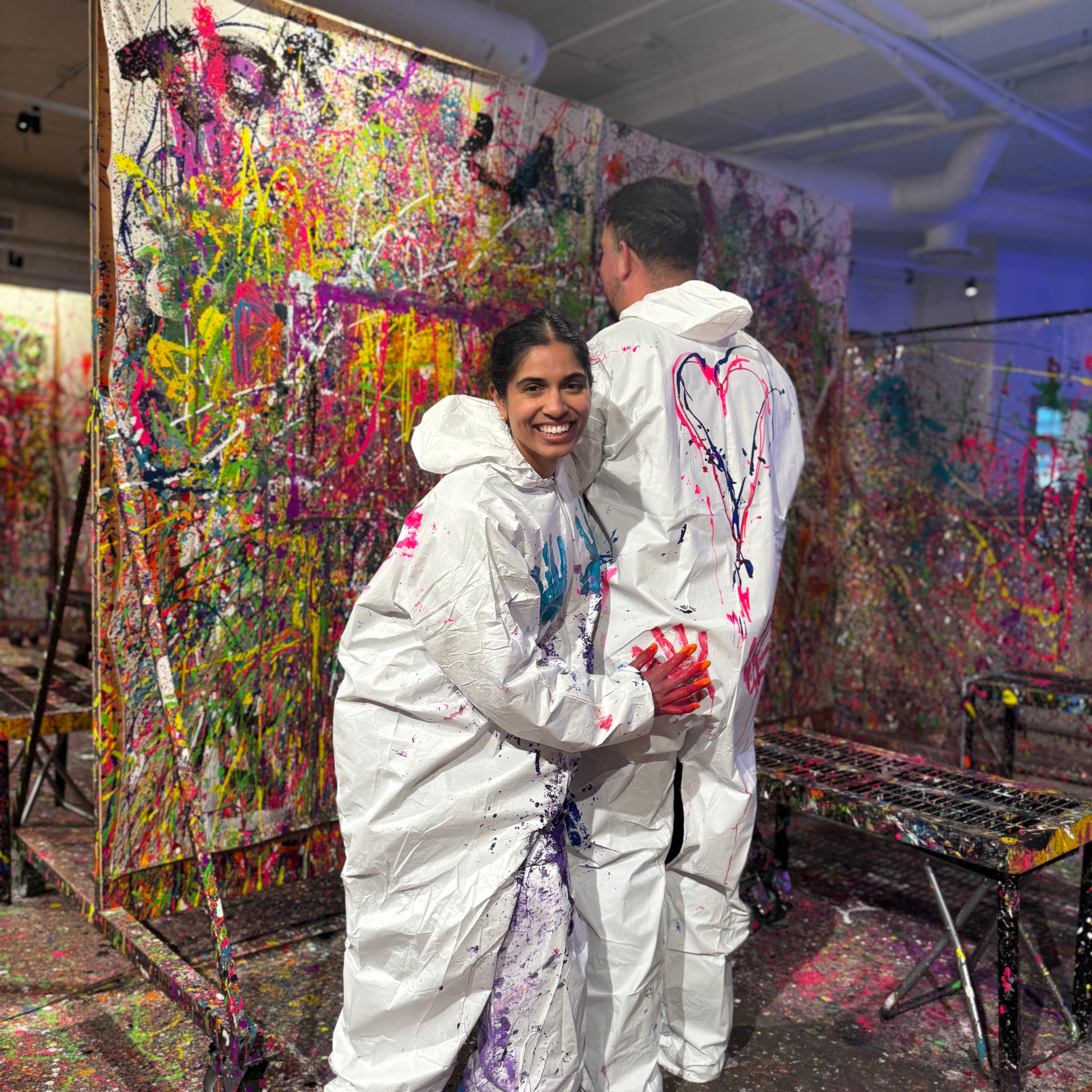 Two people in white coveralls with paint splatters, standing in a colorful splatter paint art room.