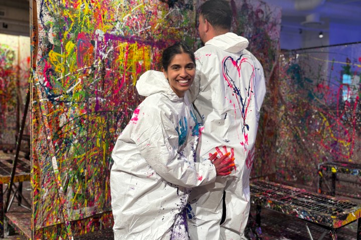Two people in white coveralls with paint splatters, standing in a colorful splatter paint art room.