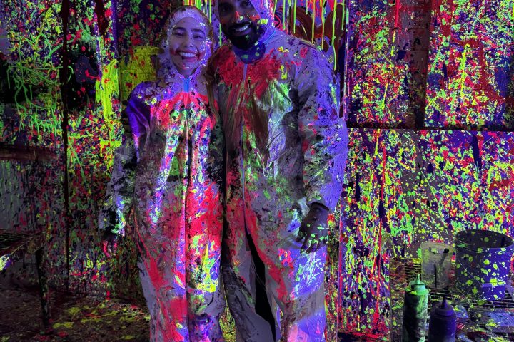 Two people in protective suits covered in neon paint, in a splatter art studio.