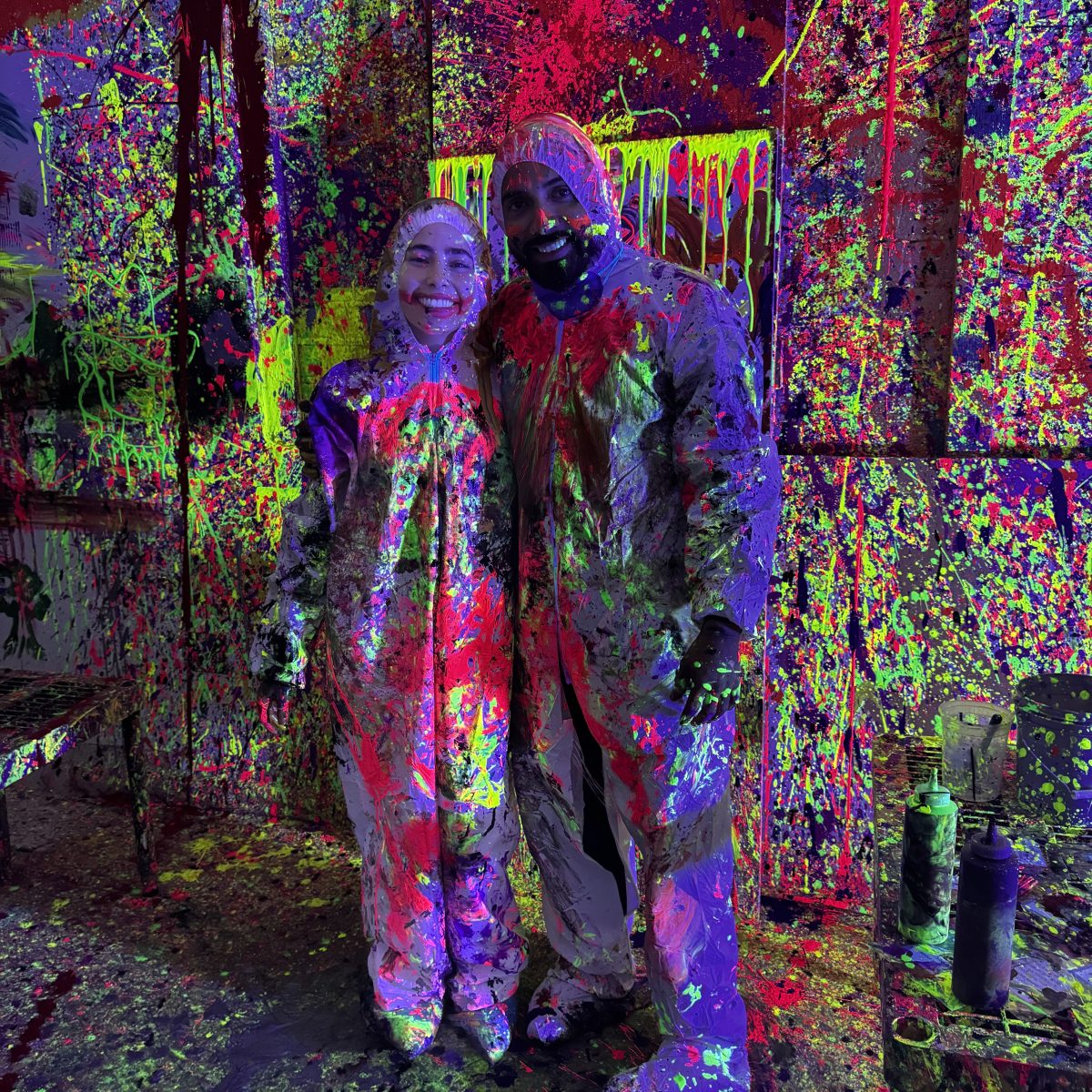 Two people in paint-splattered suits stand in a colorful, splattered room with bottles on a table.